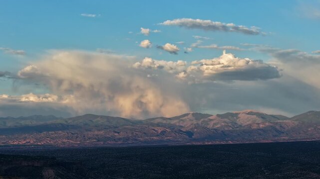 A dramatic evening timelapse capturing an isolated storm cloud releasing localized rain atop a rugged mountain range. Sweeping shadows move rapidly toward the peaks across the vast foreground valley.