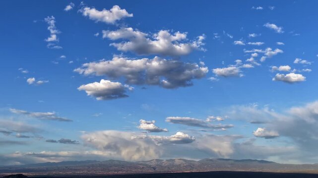 A steady timelapse capturing thick overhead clouds shifting rapidly. Evening shadows sweep across the textured ground as localized rain showers fall upon a distant, rugged mountain landscape.