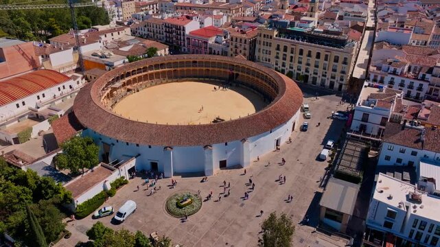 Drone footage approaching Ronda's 18th-century bullring from above, revealing its circular sand arena, terracotta roof, scattered tourists in the plaza, and dense whitewashed city surroundings.