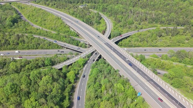 An aerial view shows a large motorway junction with cars and trucks traveling on numerous elevated roads. The intricate road network is set amidst abundant green trees during a bright day.