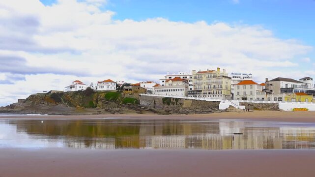 Picturesque seaside village of Praia das Macas in Sintra, Portugal, with its stunning cliffs and beautiful buildings reflected in the wet sand of the tranquil beach during low tide