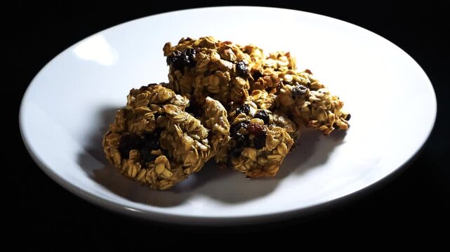 Freshly baked oatmeal raisin cookies on a white plate slowly rotating against a stark black background, highlighting their delicious, homemade texture with dramatic, moving lighting