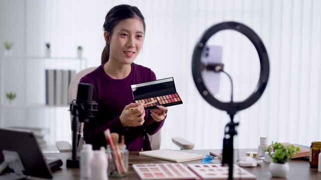 Young Asian beauty influencer presenting colorful makeup palette to camera while filming cosmetics tutorial with ring light in bright modern home studio