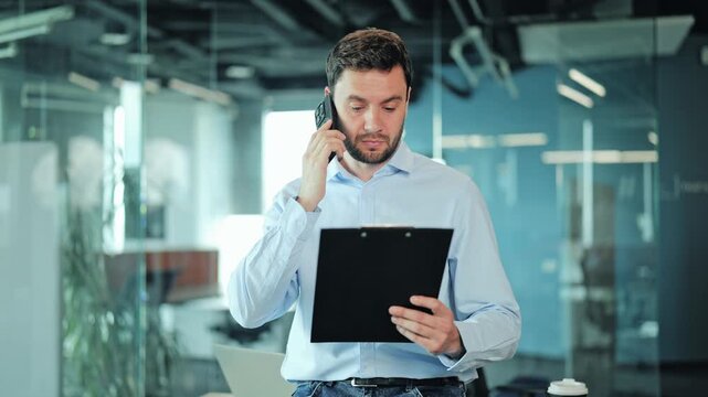 Confused corporate manager or IT specialist discussing documents on smartphone. Male employee or freelance worker looking shocked or unexpected during professional phone call in modern open office