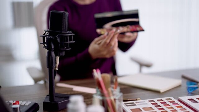 Adult woman podcaster holding makeup palette during beauty podcast recording with professional condenser microphone and cosmetic swatches on wooden desk in