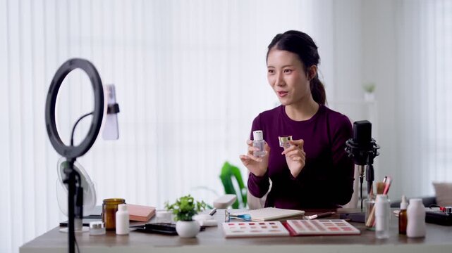 Young Asian female podcaster presenting skincare products to camera while recording beauty podcast with ring light microphone in bright home studio