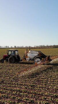 Red tractor with attached sprayer moves across a vast agricultural field under a clear blue sky, showcasing the process of crop treatment and land management