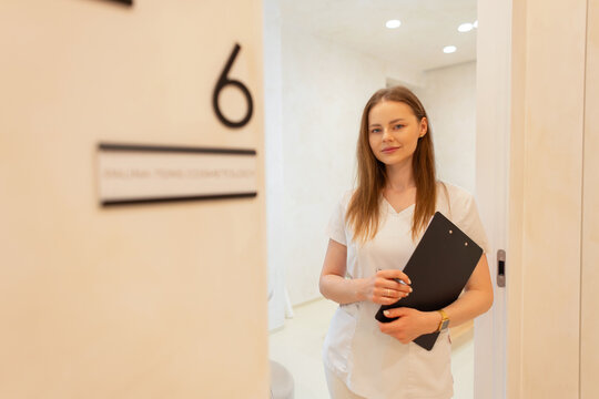 A young professional female esthetician or dermatologist in white scrubs holding a clipboard, standing in the doorway of a modern cosmetology clinic or medical office room number 6.