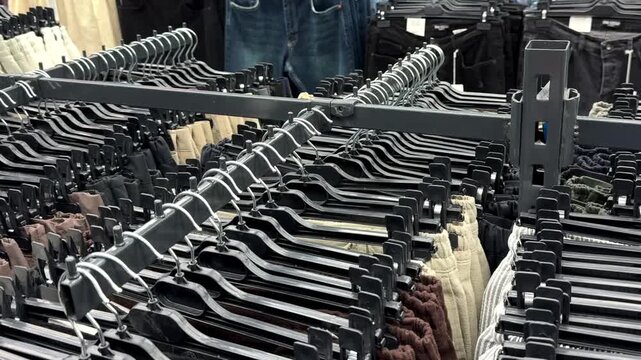 Rows of black plastic clothing hangers neatly arranged on metal racks in a retail clothing store, displaying various styles of jeans and trousers in the background.