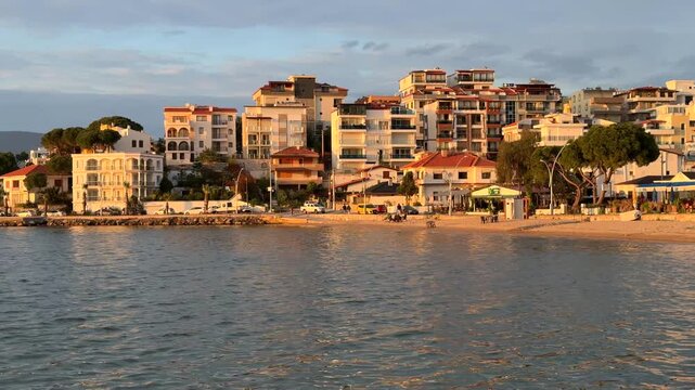 Scenic coastal view of Gulluk, Turkey, featuring white resort hotels and residential buildings along the waterfront, illuminated by the warm glow of late afternoon sun.