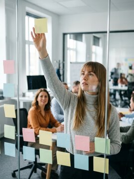 A young woman in a meeting room arranges colorful sticky notes on glass, fostering collaboration.