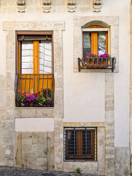 Traditional French balcony and diverse window designs on a historic building facade in Ortigia, Syracuse, Sicily, Italy. Old town stone wall with colorful potted flowers.