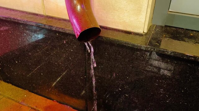 Stream of rainwater flowing out of metal building downspout at night. A steady shot of a red or dark pink drainage pipe pouring clear water onto a black polished granite tiled ground with reflections