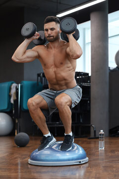 Fit and muscular man working out with bumbbells on gymnastic hemisphere bosu ball in gym.