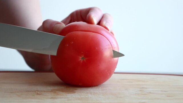 Close-up of a woman's hands slicing a large, juicy red tomato on a wooden cutting board with a sharp knife