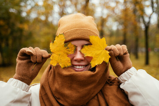 Happy young woman wrapped in scarf covering eyes with autumn leaf in public park