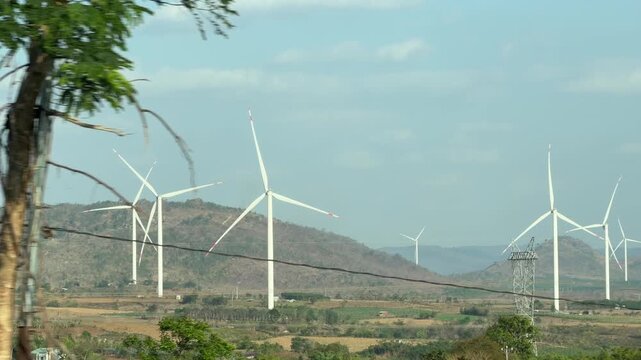 Several wind turbines rotate across open land recorded while traveling through a landscape past hills in a wide natural view under clear sky. Clean energy.