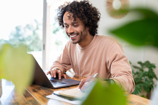 Cheerful businessman writing in diary while using laptop at home