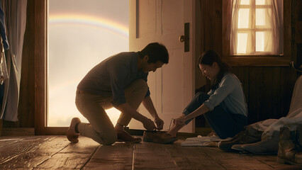 Fototapeta premium A caring young man kneeling to tie a woman's shoelaces in a cozy rustic cottage with a rainbow view.