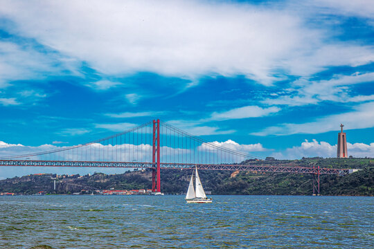PORTUGAL, LISBON - APRIL 7, 2024: Sailboat passing under the 25th April Bridge over the Tagus River,the longest suspension bridge in Europe. Statue of Christ, Santu&aacute;rio de Cristo Rei,in the background