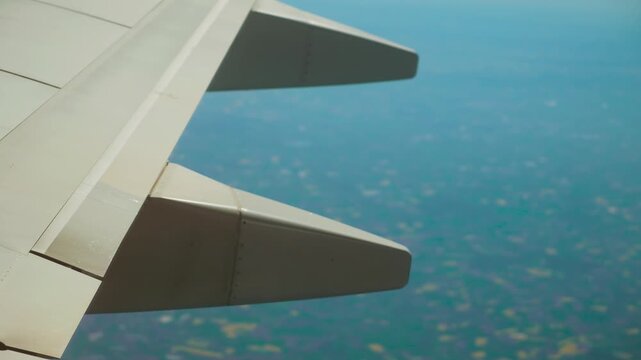 Aircraft flying above distant cityscape with visible wing and flap track fairings. Jet cruising over hazy landscape showing aerodynamic surfaces and control parts. Plane soaring through sky displaying