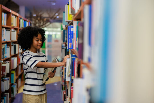 A focused young African American female student with curly natural hair stands in a bright library aisle, holding a book while carefully selecting another from a wooden bookshelf.