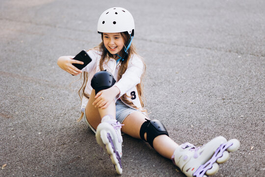 Happy girl wearing helmet and safety pads, sitting on asphalt road while taking a picture with smartphone