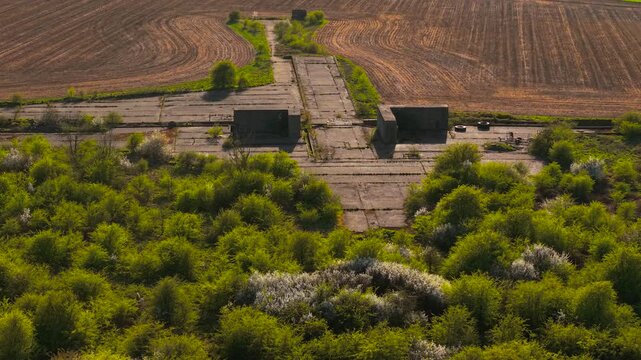 Concrete military airfield ruins geometric slabs and bunker shelters cutting through cultivated farmland patterns. Aerial drone perspective over WWII aviation site with overgrown vegetation patches