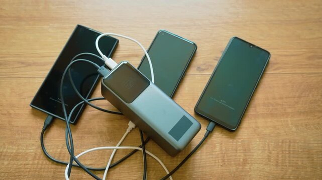 Power bank charging three smartphones simultaneously using multiple cables portable charger powering mobile devices. External battery delivering energy to several phones at once in active charging