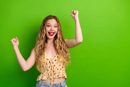 Happy young woman with blonde hair in a floral top raises arms in joy against a bright green background