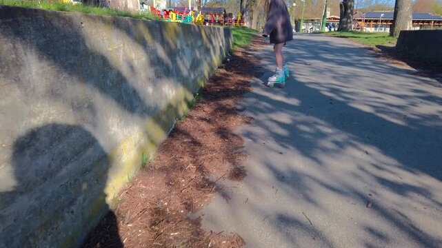 Outdoor static wide shot captures a young girl in inline skates navigating a steep paved incline past a concrete retaining wall next to trees and a park playground on a bright sunny day.