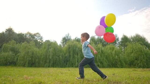 Boy runs with colorful balloons across meadow outdoors. Happy child plays with balloon bunch in park. Kid running through meadow field. Boy holds balloons while running. Child enjoys balloon fun.