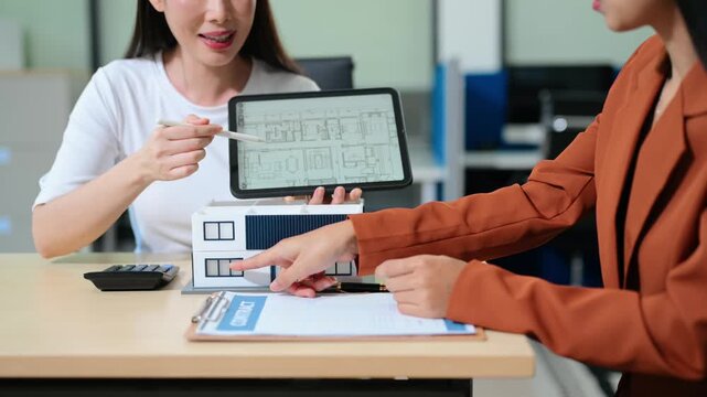 Professional architects discuss house blueprints on a digital tablet with a model home and contract on the table.
