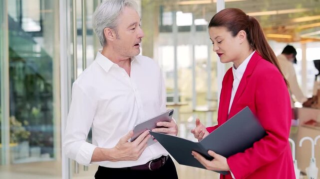 Office meeting with senior man and woman discussing documents in modern workplace lobby, business conversation in glass office interior