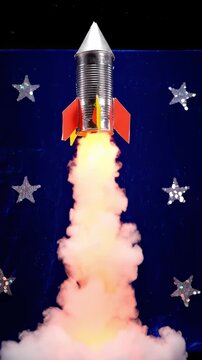 Toy rocket made from a tin can launches upwards with smoke and flames. Handmade diorama features tangible textures of fabric, metal, and cotton wool.