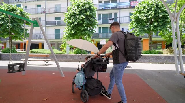 Young dad with a backpack pushing his toddler son in a baby stroller through a city park, stopping to carefully attach an umbrella to provide shade and protection from the sun