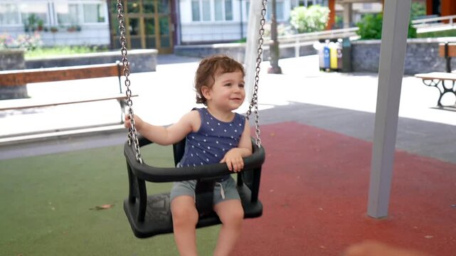 Happy little boy with curly hair smiling and laughing while his loving father pushes him on a swing set at an outdoor playground, creating a beautiful family moment during a sunny day