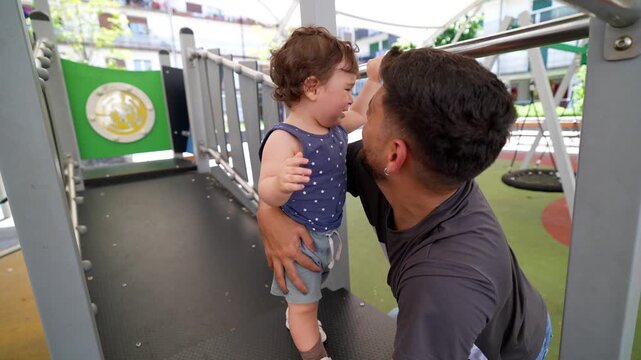 Affectionate young father tenderly kissing and comforting his upset toddler son while playing together at an outdoor public park, showcasing a beautiful moment of paternal love and care