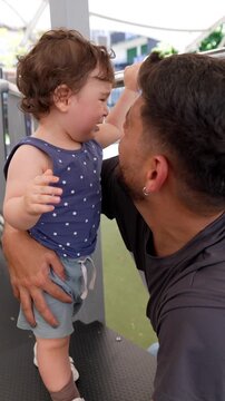 Affectionate father holding and consoling his little toddler son who is crying at an outdoor playground, showing a beautiful moment of paternal love, comfort, and emotional connection