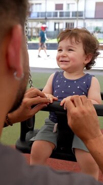 Adorable little boy with curly hair smiling and laughing joyfully while his loving father gently pushes him on a swing set during a sunny day at the park, enjoying quality family time together