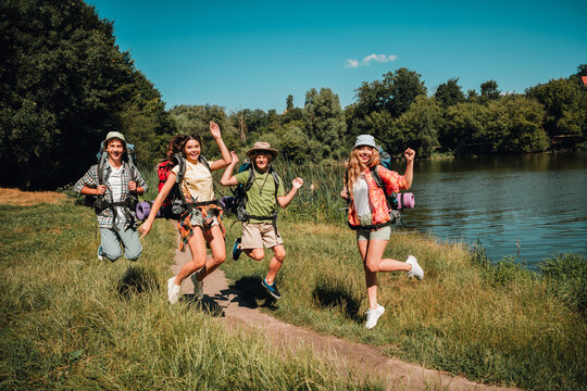 Friends hiking and jumping with backpacks by a lake on a sunny summer day &mdash; joyful group of teenagers enjoying outdoor adventure and nature