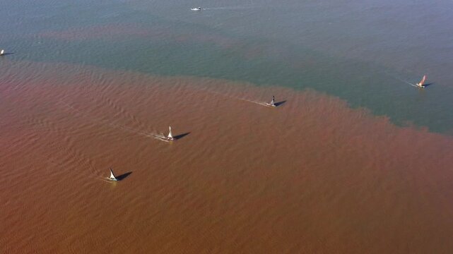 High angle drone footage of a fleet of traditional Malagasy fishing pirogues sailing across a stark water sediment line in Mahajanga, Madagascar.