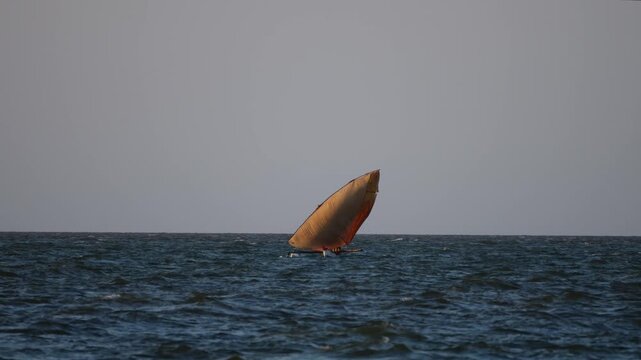 Long lens shot of a traditional Malagasy dhow boat with a large orange sail catching the wind on the choppy sea during sunset in Mahajanga, Madagascar.