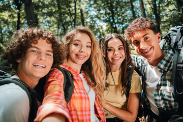 Obraz premium Four smiling teenage friends taking a group selfie while hiking in a sunny forest with backpacks and casual summer clothes