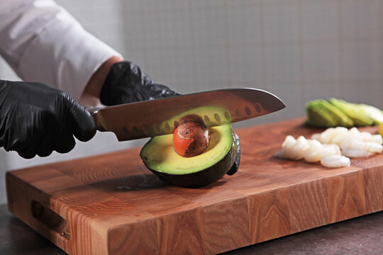 Close-up of chef slicing an avocado with knife on cutting board. Cook cuts avocado. Ripe avocado in hands of chef. Healthy and low-calorie fruit. Hands in protective gloves. Concept of cooking at home