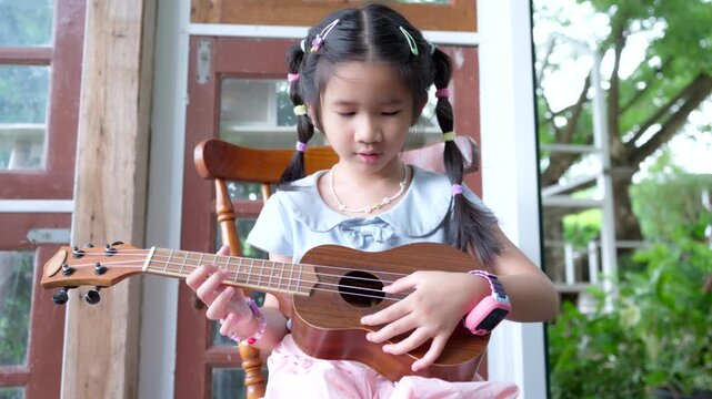 Cute little Asian girl playing ukulele happily while sitting on a wooden chair, childhood learning and music education concept.