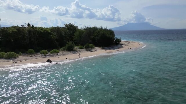 Aerial view of a girl runs on the beach as the camera pans out to reveal the clear turqoise waters and backdrop, Tabuhan Island off the coast of Java and Bali