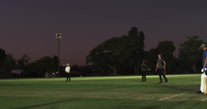Asian man running and bowling ball testing teenage batsman on pitch, floodlights, copy space