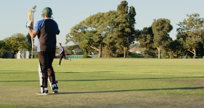 Walking diverse male teammates carrying wooden bats across cricket ground, with helmets, gloves