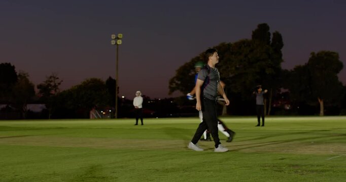 Diverse teammates bowling on floodlit cricket pitch scratching head, adjusting hat before run-up